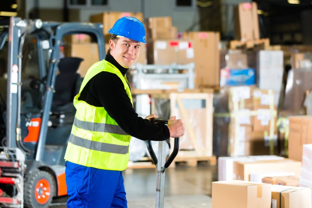 Warehouseman in protective vest pulls a mover with packages and boxes at warehouse of freight forwarding company- a forklift is in Background